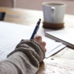 Person writing on brown wooden table near white ceramic mug and notepad