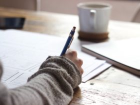 Person writing on brown wooden table near white ceramic mug and notepad