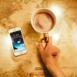 Person's hand holding a mug with coffee next to an iphone and on top of a world map