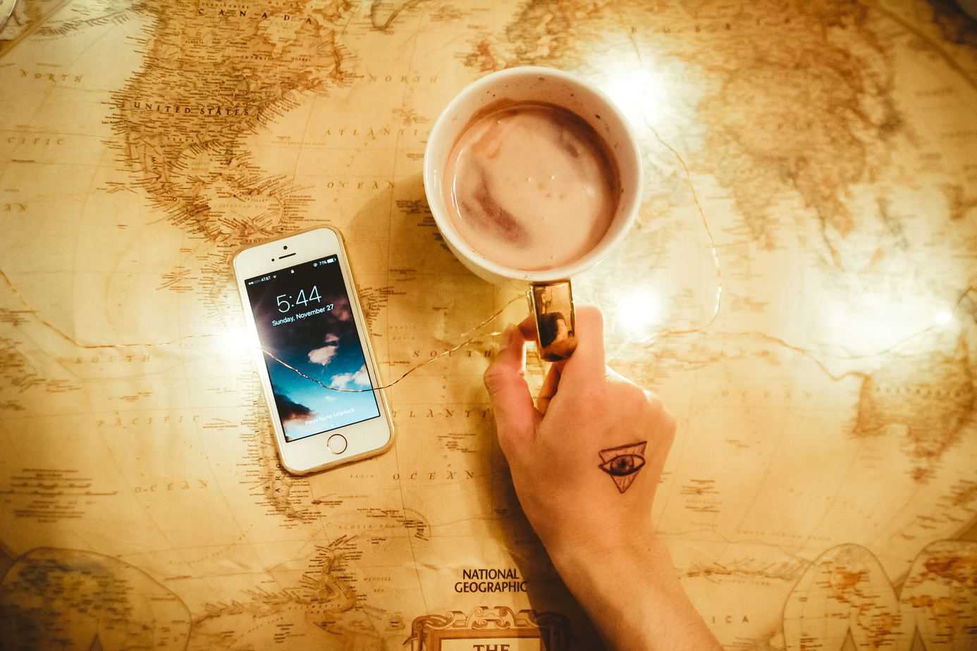 Person's hand holding a mug with coffee next to an iphone and on top of a world map