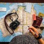 Person holding a pen on top of an Australian map next to passport, money, sunglasses and a camera
