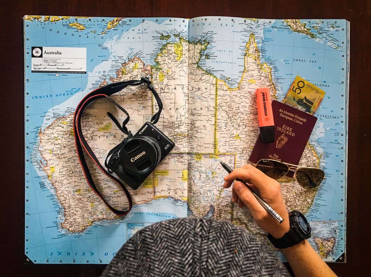 Person holding a pen on top of an Australian map next to passport, money, sunglasses and a camera