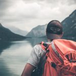 Man with red hiking backpack facing body of water and mountains at daytime