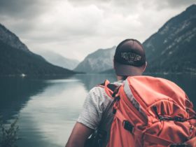 Man with red hiking backpack facing body of water and mountains at daytime