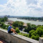 Cameron sitting on a concrete seat in front of Serbia while wearing sunglasses and a baseball cap