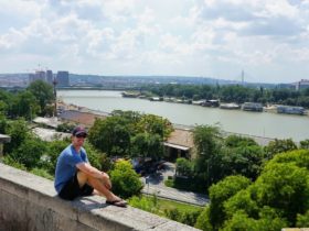 Cameron sitting on a concrete seat in front of Serbia while wearing sunglasses and a baseball cap