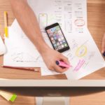 Person writing in white paper with colors and surrounded by a computer and a smartphone