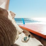 Silver framed sunglasses next to surfboard near ocean