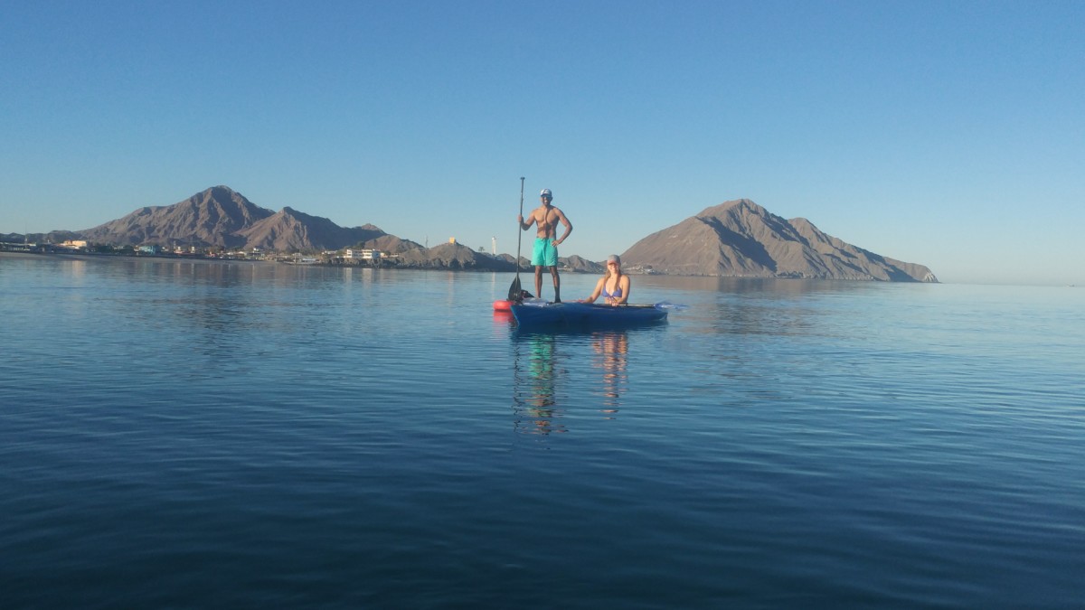 Julie sits in a blue kayak on a lake while Reet stands on a red stand-up paddboard