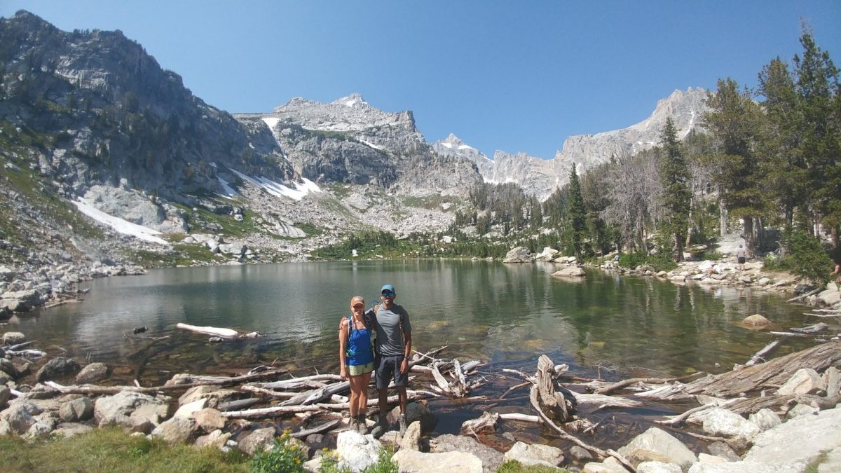 Julie and Reet Singh stand in front on an alpine lake on a sunny day with sparse trees and some higher mountain peaks behind them