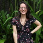 Deya smiles with her hand on her hip while wearing a dress and glasses, posing in front of topical green plants