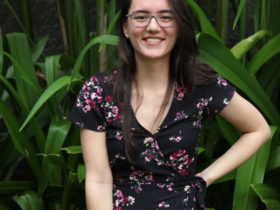 Deya smiles with her hand on her hip while wearing a dress and glasses, posing in front of topical green plants