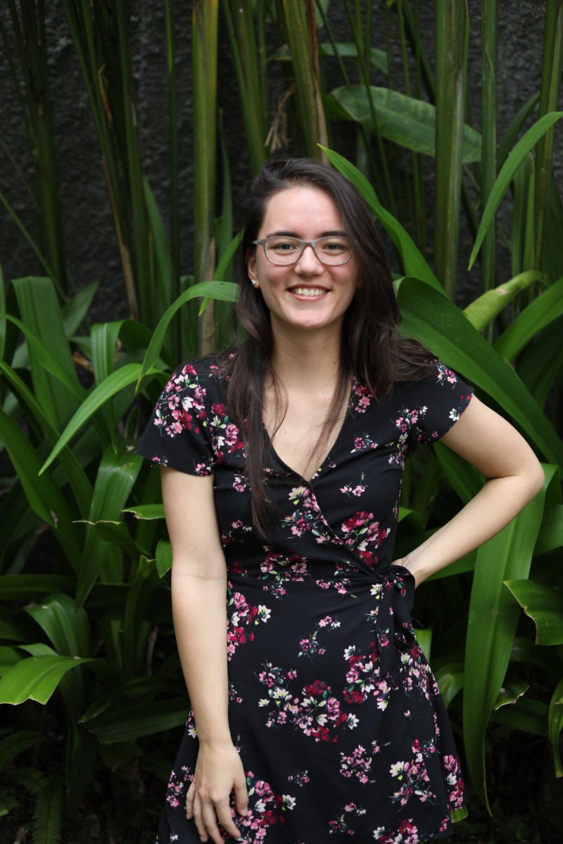 Deya smiles with her hand on her hip while wearing a dress and glasses, posing in front of topical green plants