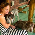 Jane Thomas sits on a front porch with one dog cuddling her and another dog sitting at her lap