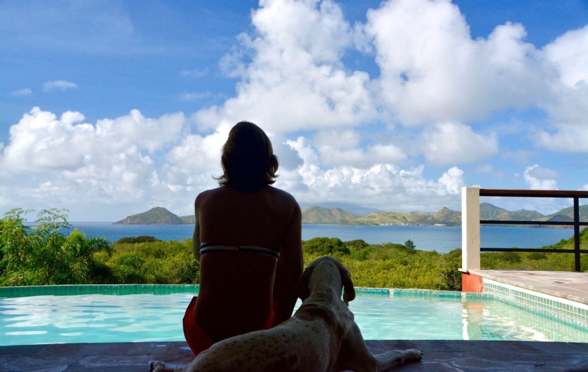 Jane Thomas sits next to a pool with her back to the camera while a dog lays beside her overlooking the ocean and a mountain