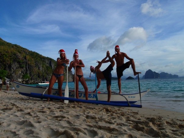 Jane Thomas stands with four friends on a beach in the Philippines wearing santa hats and standing next to an outrigger canoe