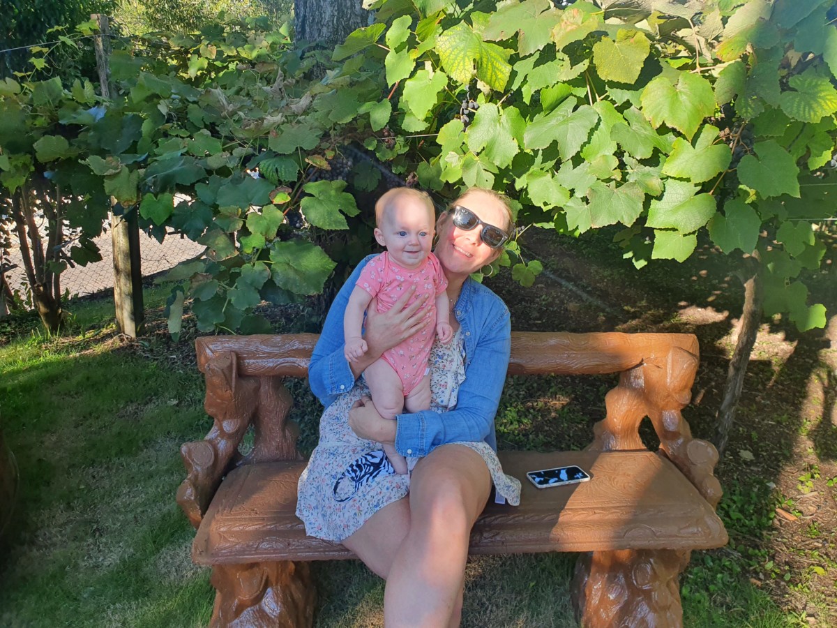 Katie Davis sits on a park bench on a sunny day with her daughter standing in her lap