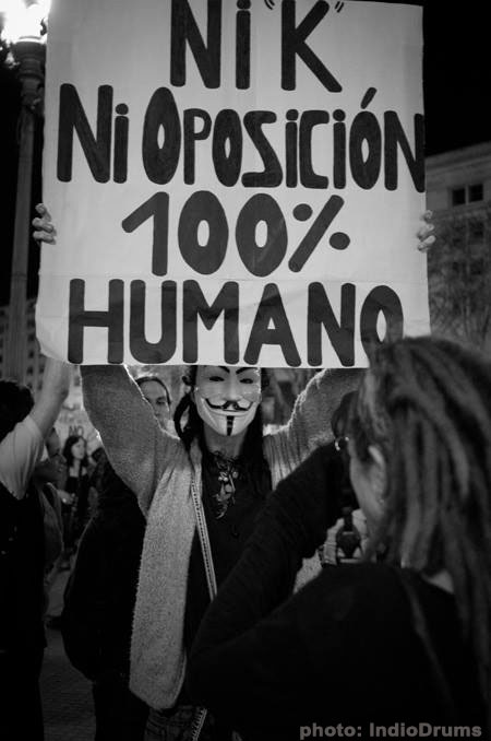 A black and white image shows Ande Wanderer taking a photo of a protester in Bueno aires who is wearing a guy Fawkes mask and holding a sign that says ni k ni oposicion 100% humano