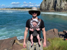 Brian Davis in Torres wearing his daughter on a front pack with the ocean and cliffs behind them