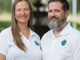 Sean and Julie chickery wear matching white polo shirts, slightly facing once another and smiling with a blurry background behind them