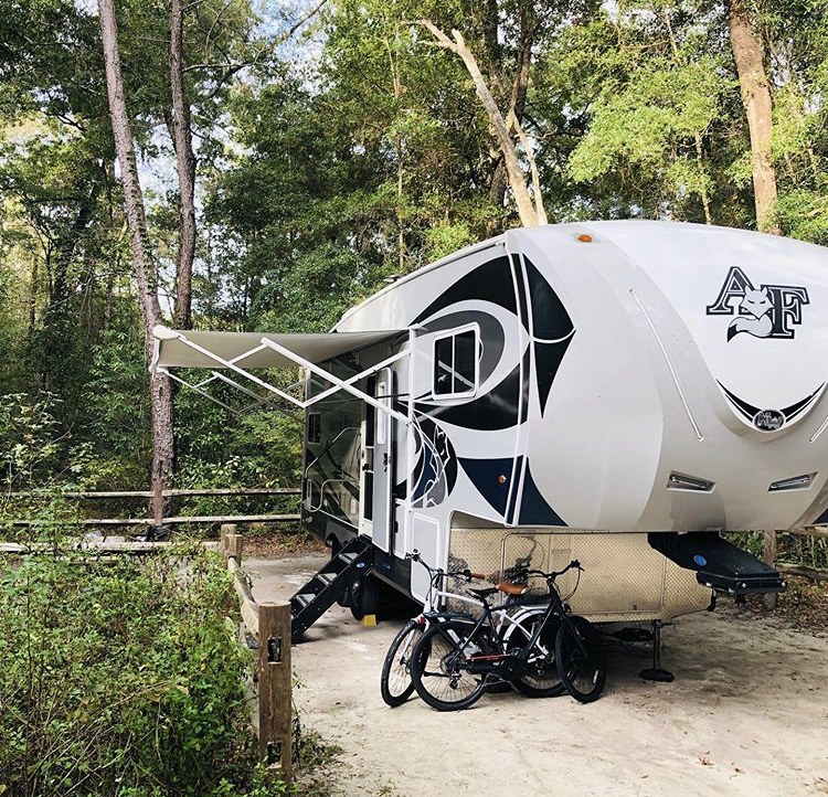 an RV sits parked in a camp spot with the awning out and two bicycles sitting in front of the RV