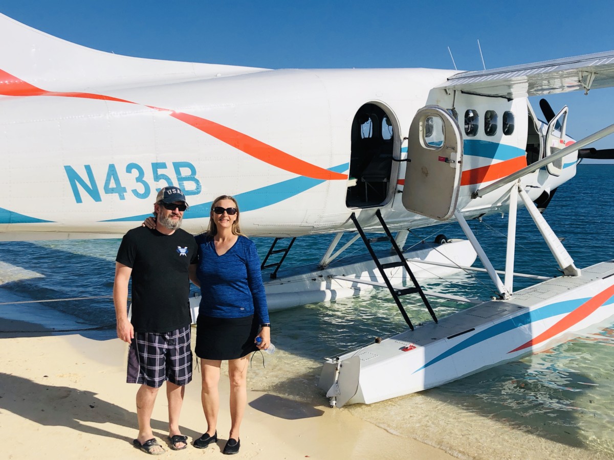 Sean and Julie chickery stand in the sand next to a sea plane that is floating in the water nearby