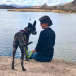 Amy Burkert fits on a slight grassy slope with a dog standing next to her and a large lake in front of them