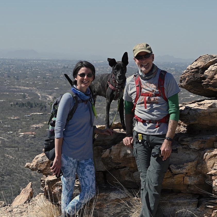Amy Burkert and her huband stand wearing backpacks on some rocks that overlook a canyon while their black dog stands behind them on some rocks
