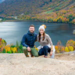 Mark and Kristen Morgan squat on a rock with a large lake behind them surrounded by trees with vibrant fall colors