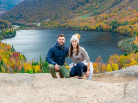 Mark and Kristen Morgan squat on a rock with a large lake behind them surrounded by trees with vibrant fall colors