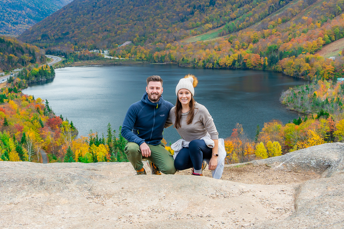 Mark and Kristen Morgan squat on a rock with a large lake behind them surrounded by trees with vibrant fall colors