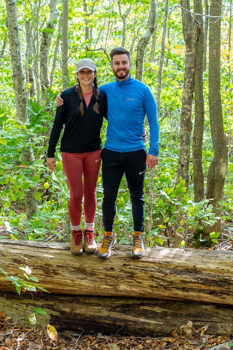 Mark and Kristen Morgan stand on a fallen tree trunk while wearing hiking clothes in a dense bright green forest