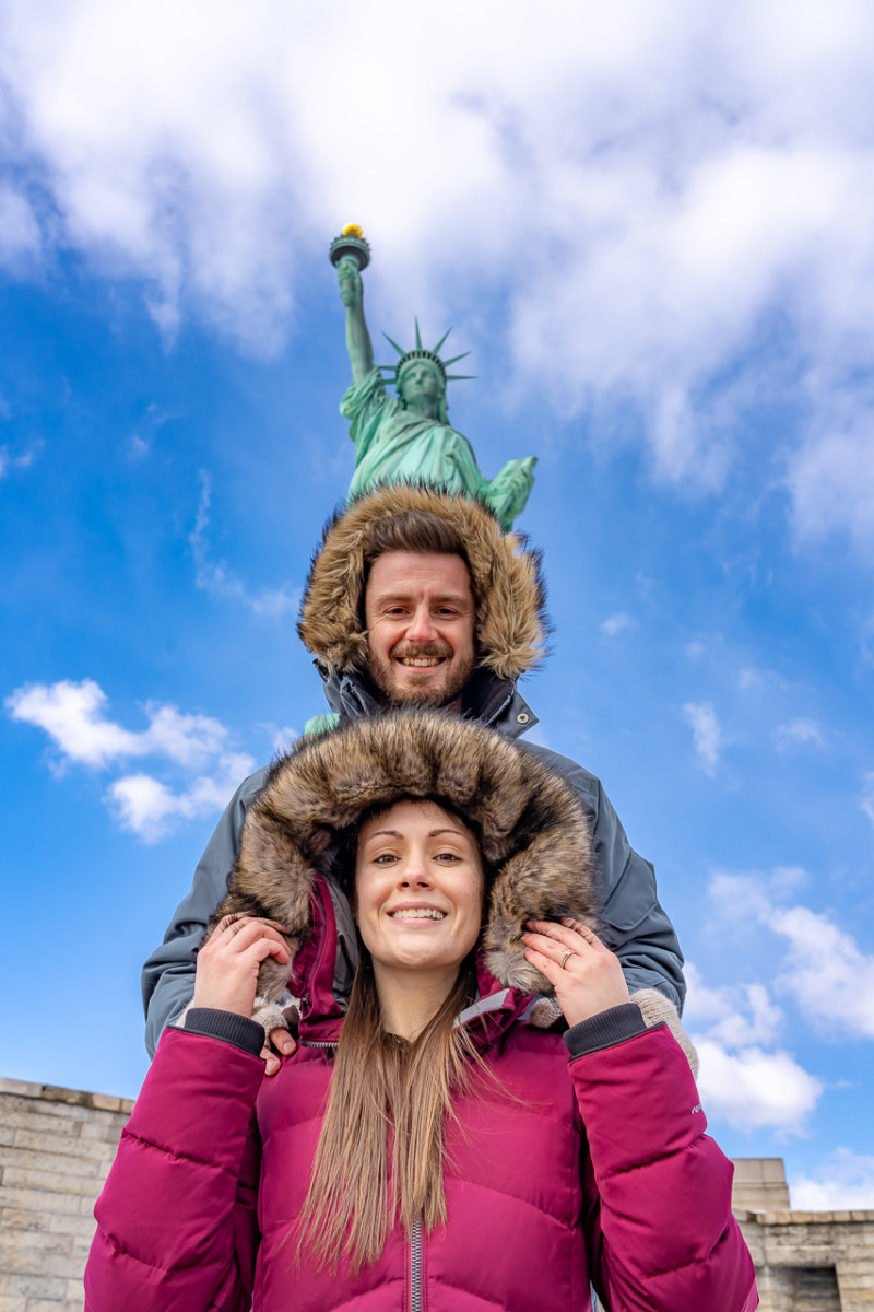 Kristen Morgan stands in front of Mark Morgan who stands in front of the statue of liberty on a cold, sunny day