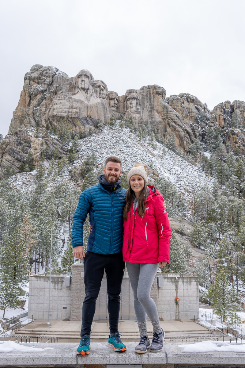 Mark and Kristen Morgan stand with their arm around each other in front of Mt. Rushmore with some snow on the ground