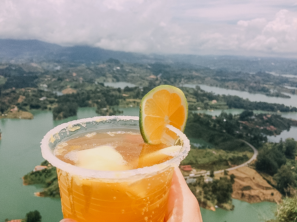 Anny Wooldridge holding a plastic cup with an orange drink in front of the city of Guatapé in Columbia