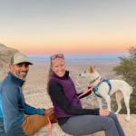 Julie and Reet Singh sit atop a rock with their small dog smiling at the camera with a multicolored sunset in the background