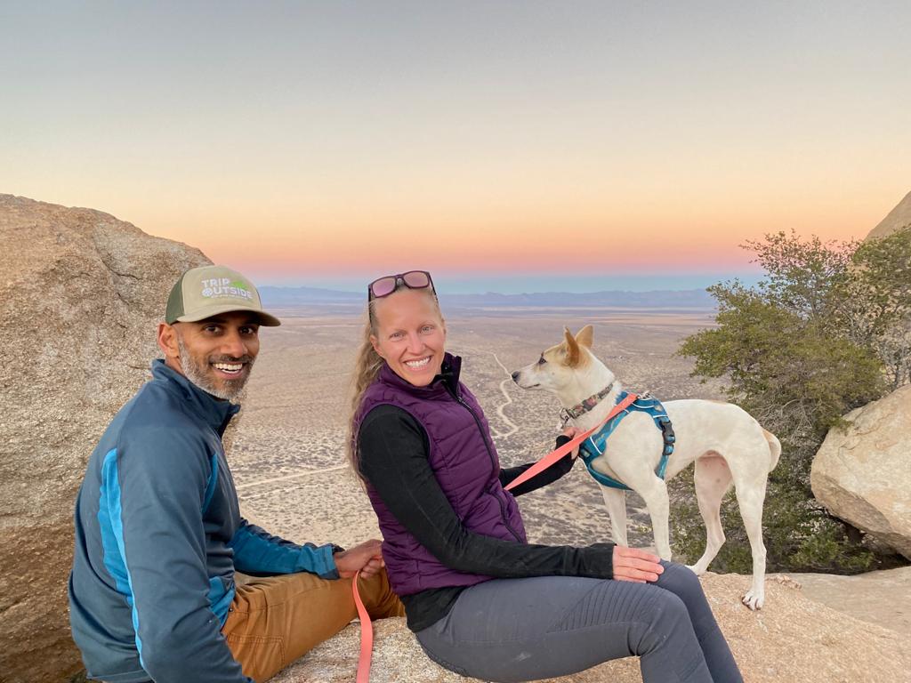 Julie and Reet Singh sit atop a rock with their small dog smiling at the camera with a multicolored sunset in the background