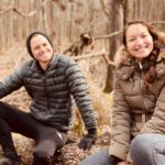 Amy Suto and Kyle Cords sit on a log in a brown forest while wearing puffy coats