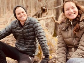 Amy Suto and Kyle Cords sit on a log in a brown forest while wearing puffy coats