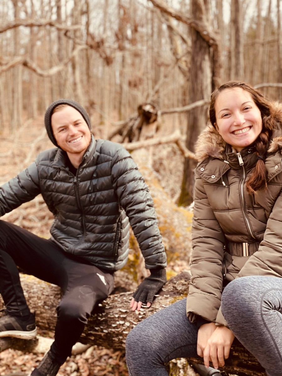 Amy Suto and Kyle Cords sit on a log in a brown forest while wearing puffy coats