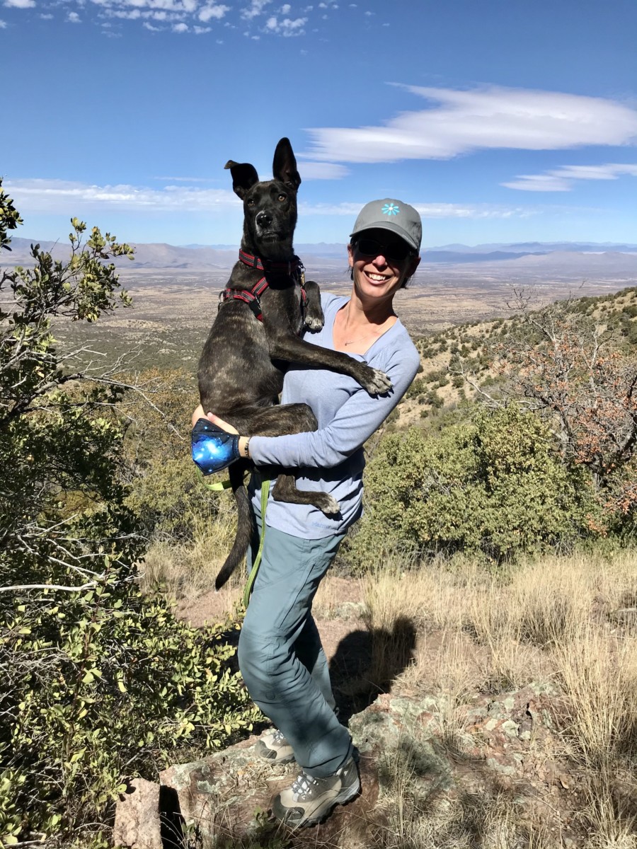 Amy Burkert wears hiking attire and a baseball hat in a desert environment while holding up a dark colored dog