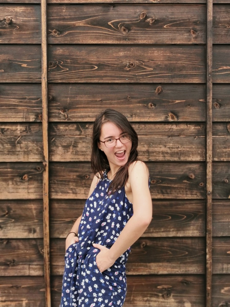 Deya stands with her hands in her dress pockets in front of a wooden panel wall while making a silly face