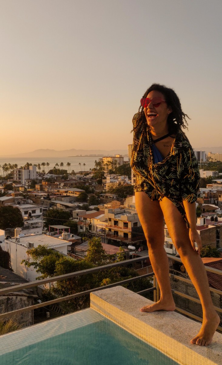 Felly days stands on the ledge of a pool at sunset with a large metropolitan city behind her