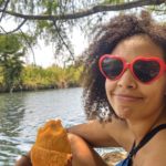 Felly day sits by a lake with a peeled mango in her hand while wearing red, heart-shaped sunglasses