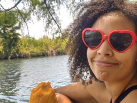 Felly day sits by a lake with a peeled mango in her hand while wearing red, heart-shaped sunglasses