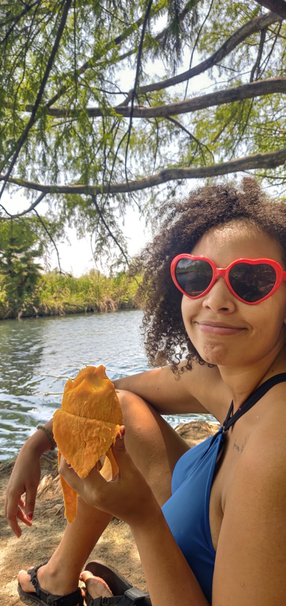 Felly day sits by a lake with a peeled mango in her hand while wearing red, heart-shaped sunglasses