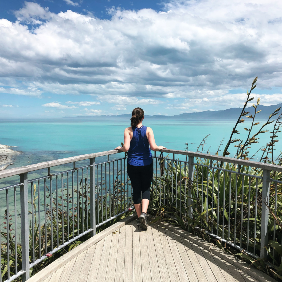 Nina Clapperton stands at the corner of a chest-high fence overlooking a turquoise ocean with a slightly cloudy but blue sky above