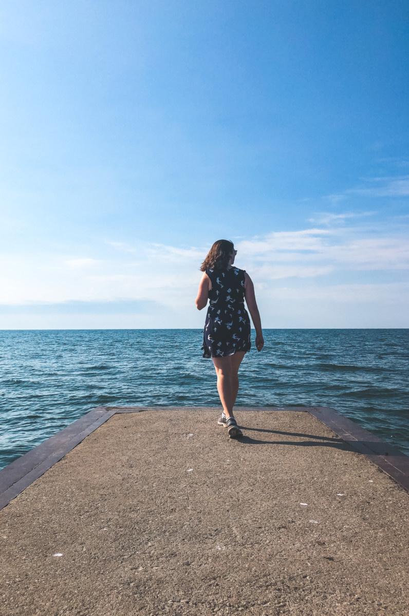 Nina Clapperton walks to the end of a concrete pier with the blue ocean in front of her while the sun is shining