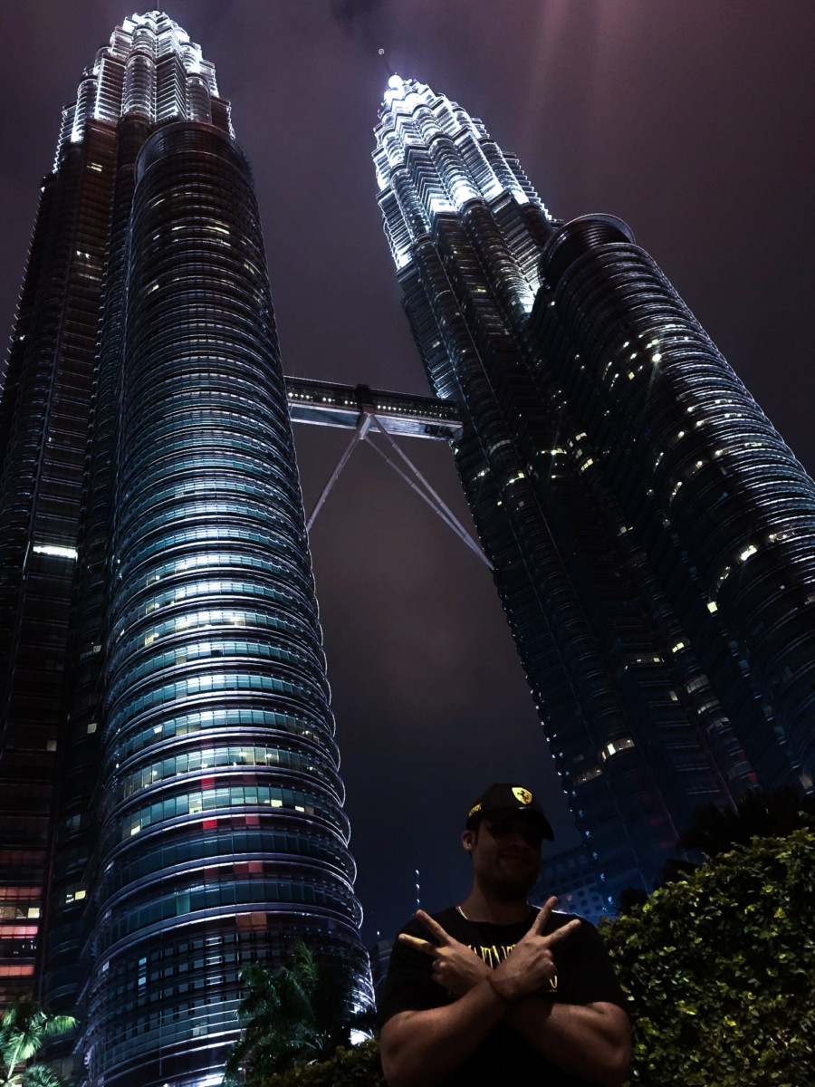 Phillip Lew standing at the base of the Petronas Towers in Kuala Lumpur at night with his arms crossed and fingers in a peace signs