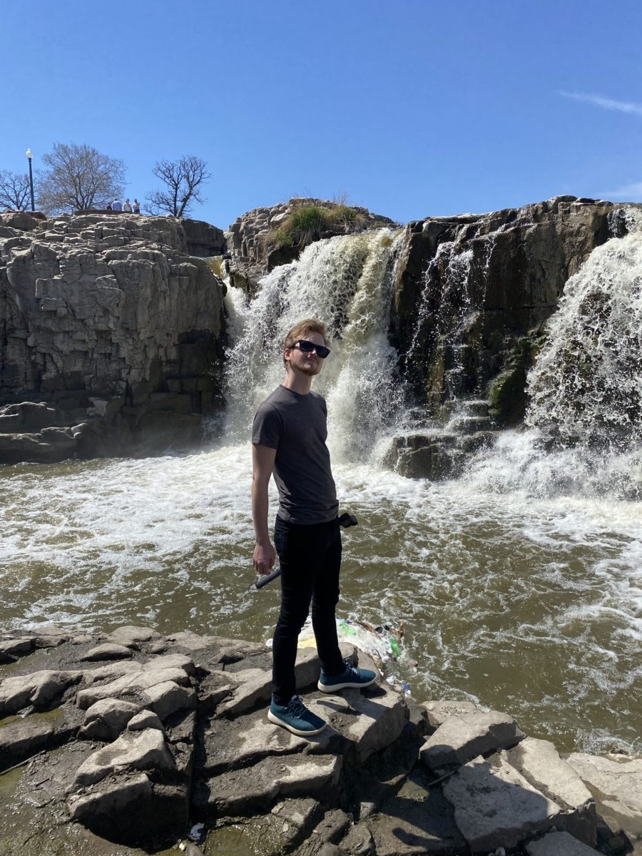 Kyle cords stands on a rock outcropping in front of a fast-flowing waterpool while wearing sunglasses and smiling on a sunny day